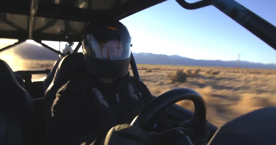 A person wearing a helmet and racing gear is driving an offroad vehicle through a desert landscape with mountains in the background during sunset