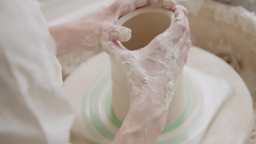 A person shapes a clay pot on a pottery wheel hands covered in wet clay focusing on forming the pots surface
