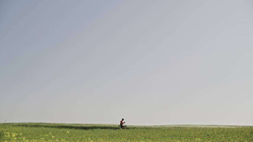 Reveal of a lone cyclist in a large lush green rural landscape under a blue sky getting on his bike and starting pedaling