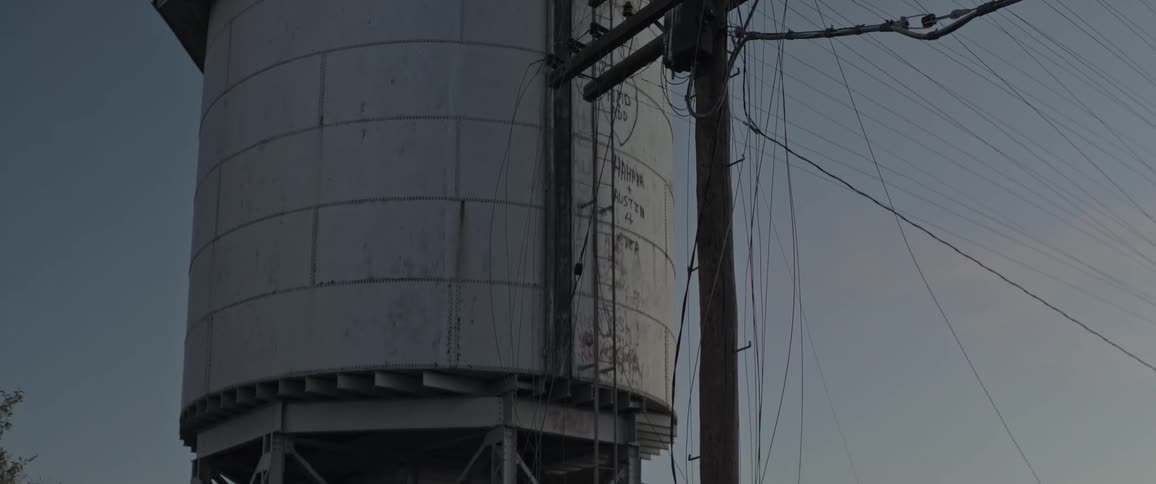 A large cylindrical water tower stands next to a utility pole with multiple crossarms and insulators in the countryside at dusk