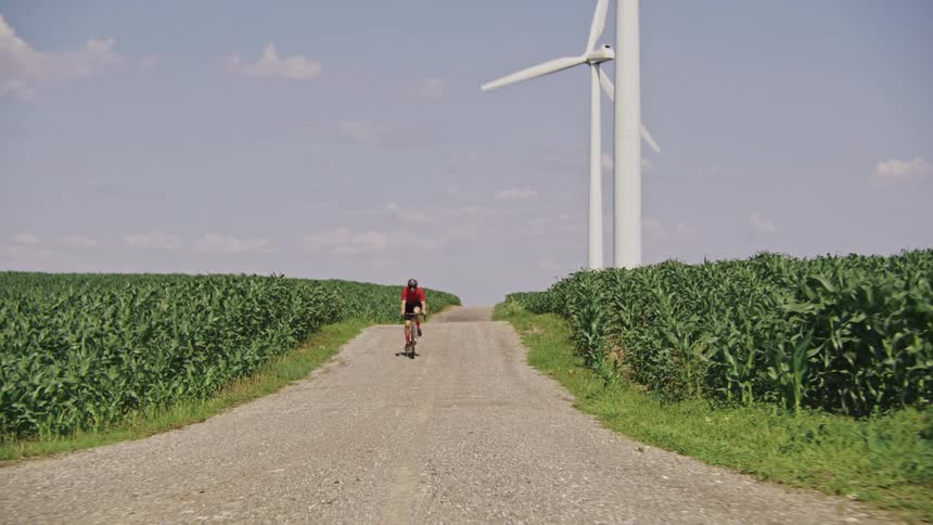 A gravel cyclist passing by on a gravel road surrounded by corn fields with wind turbines in the background under a clear blue sky