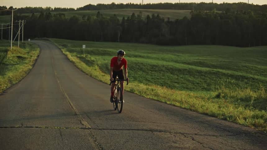 A tired cyclist riding up a paved road at sunset surrounded by green fields and hills in the background