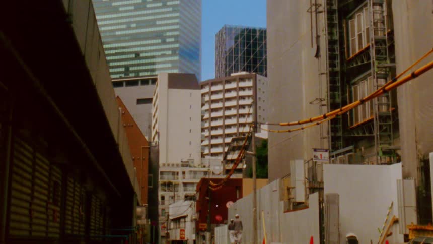 A narrow urban alleyway is flanked by tall buildings under construction with visible scaffolding and construction materials