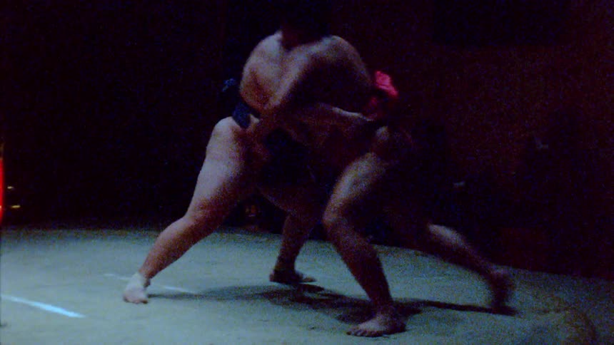 A couple of sumo wrestlers engaged in a match on a dimly lit ring