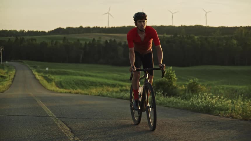 A male road cyclist at sunset riding up a hill surrounded by lush green vegetation, wind turbines and rolling hills and in a rural landscape