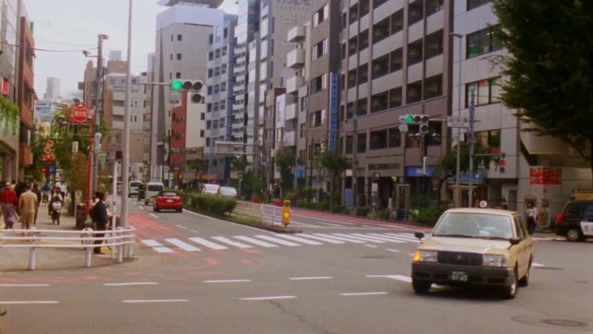 Traffic flows through a busy intersection in a japanese city with pedestrians crossing the street amidst a bustling cityscape