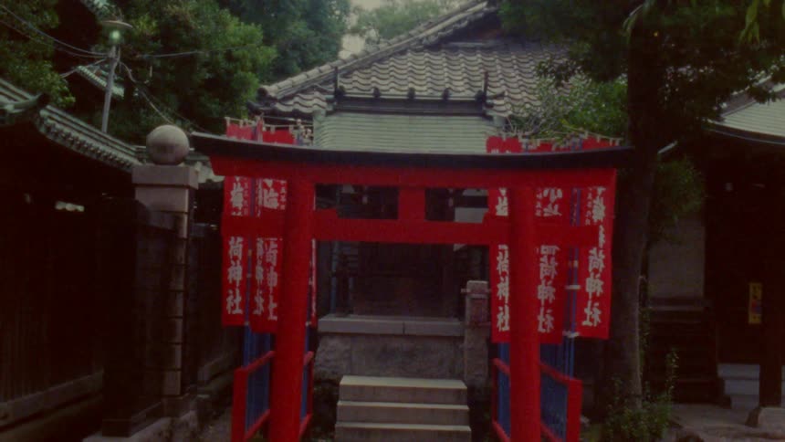 The image shows a traditional japanese shrine entrance with a red torii gate stone steps and surrounding greenery leading to a building with a tiled roof