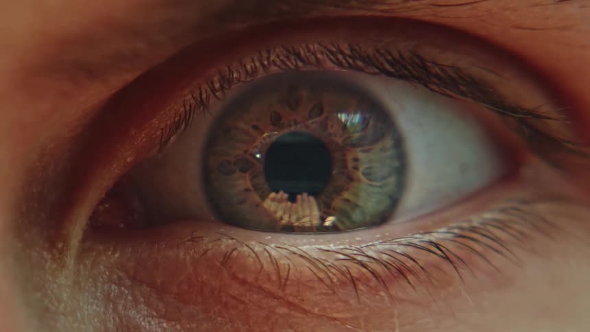 A close up image of a human eye with an iris rapidly expanding and reflections of hands visible in the eye