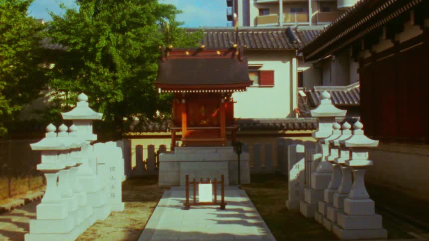 Stone lantern statues lead to a small shrine near buildings on a sunny day