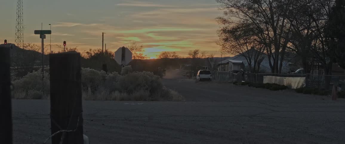 A rural scene at sunset with a dirt road leading to a small settlement of houses and a parked car framed by leafless trees and utility poles with a colorful sunset sky