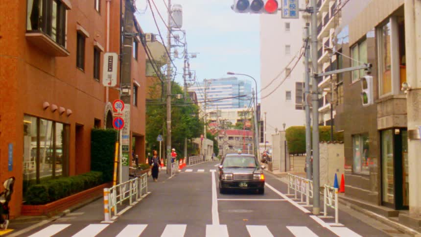 A taxi cab drives down a quiet residential street in japan lined with lowrise buildings and a crosswalk with a red light