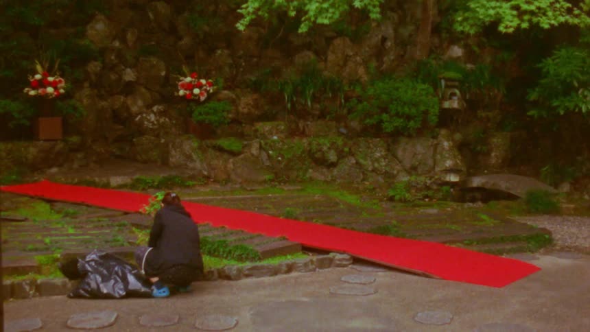 A person kneels on a stone path next to a red carpet leading to a rocky mosscovered area with flower arrangements