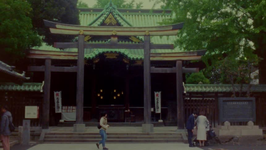 Traditional japanese shrine with a torii gate in the foreground a greentiled roof and people walking and standing near the entrance