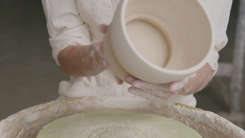 A person is inspecting a freshly crafted ceramic pot above a pottery wheel with hands covered in clay