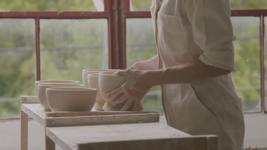 A potter inspects a newly formed ceramic bowl in a workshop filled with natural light