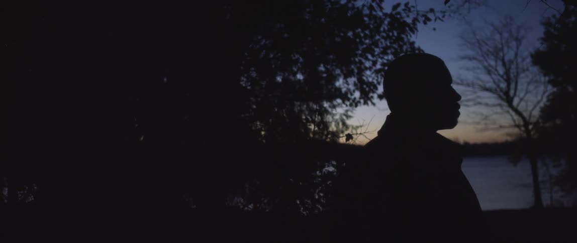 A silhouetted man stands near a lake at dusk