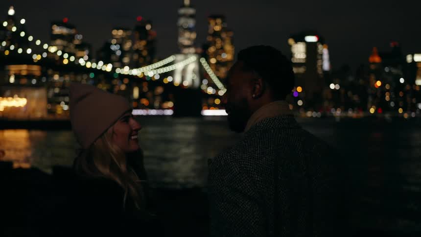 Black man and white blonde woman walking and hugging outdoors at night next to a river with a metropolis in the background