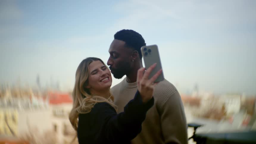 A young couple stands on a balcony taking selfies wiht a city under a blue sky in the background