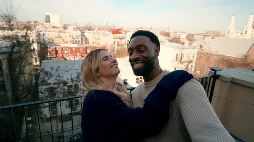 A smiling diverse couple takes a selfie on a rooftop with a city skyline in the background and the woman hugs and kisses the man