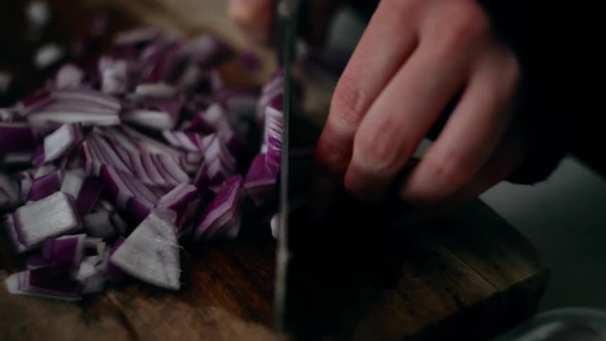 A hand holds a knife and slices a red onion on a wooden cutting board