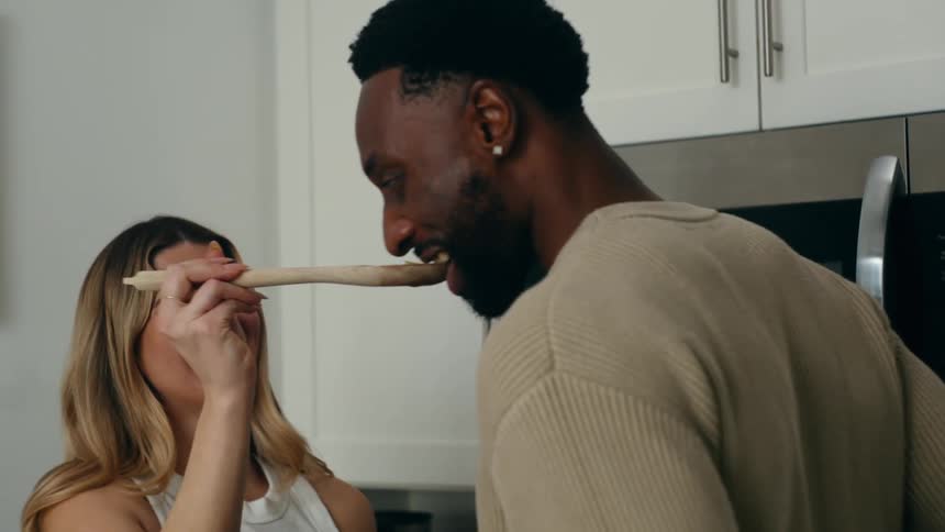 Man smiles while tasting food offered on a wooden spoon by blonde female cook