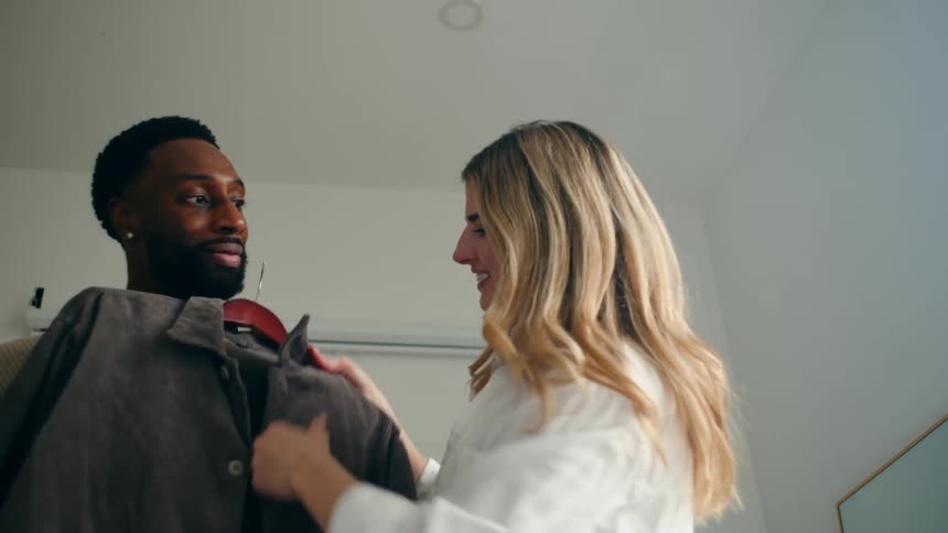 White blonde woman in white clothes trying different jackets over the body of a black man while both laugh and talk indoors during day time