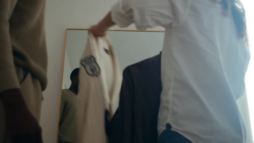A woman holds up clothes to a man in a room with a mirror