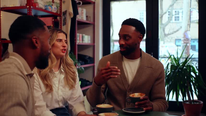 Three people sit at a table in a caf engaging in conversation while holding cups of coffee with shelves in the background