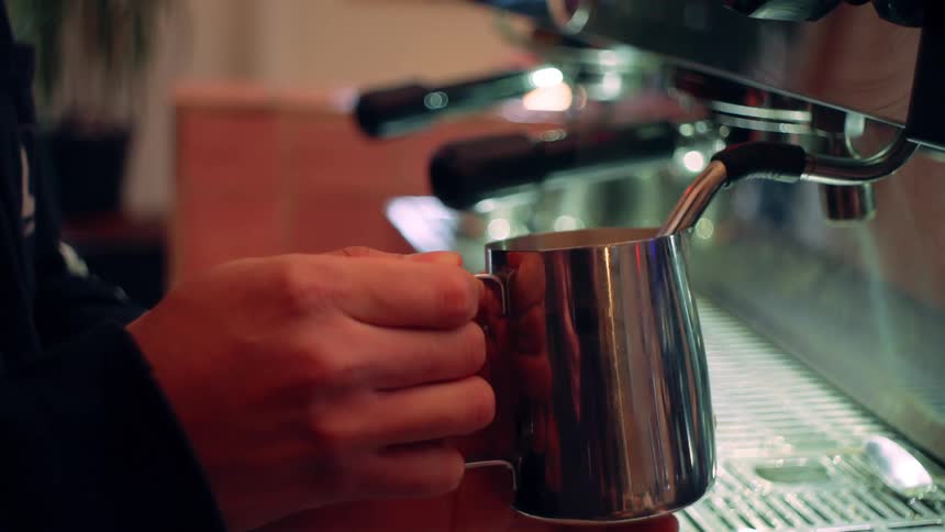 A baristas hand holds a stainless steel pitcher under an espresso machine spout