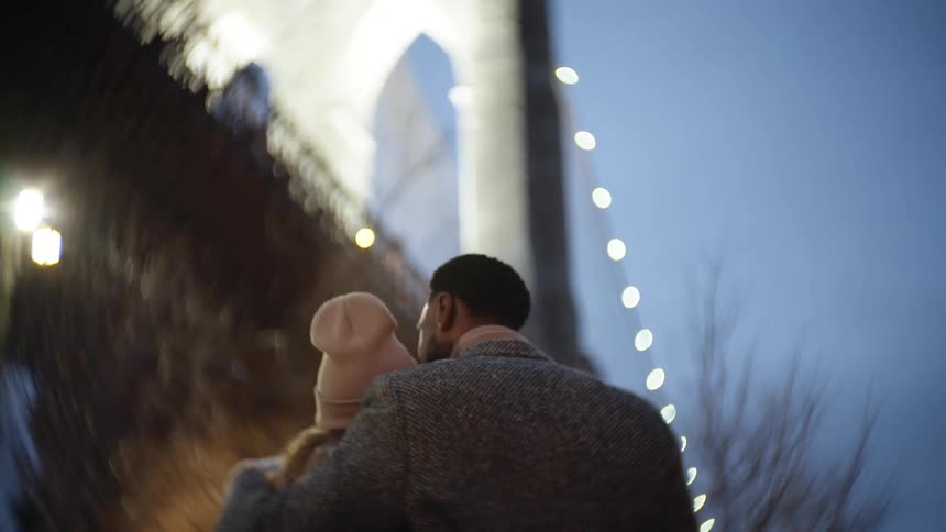 A couple is seen from behind embracing as they walk towards the iconic arches of a bridge at twilight with a prominent bokeh effect around the edges