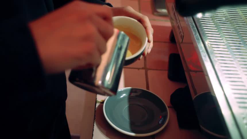 Person standing up indoors next to a counter preparing a cup of coffee latte