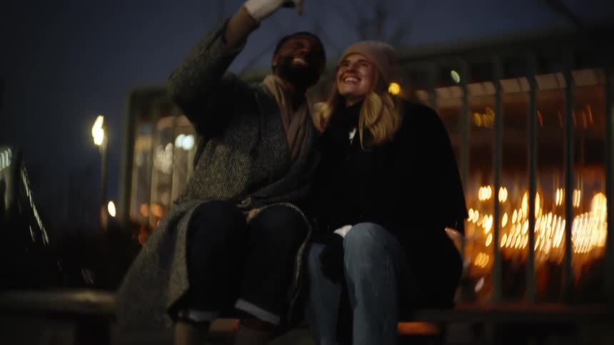 Happy couple taking selfie with a smartphone while sitting outdoors on a bench in the evening