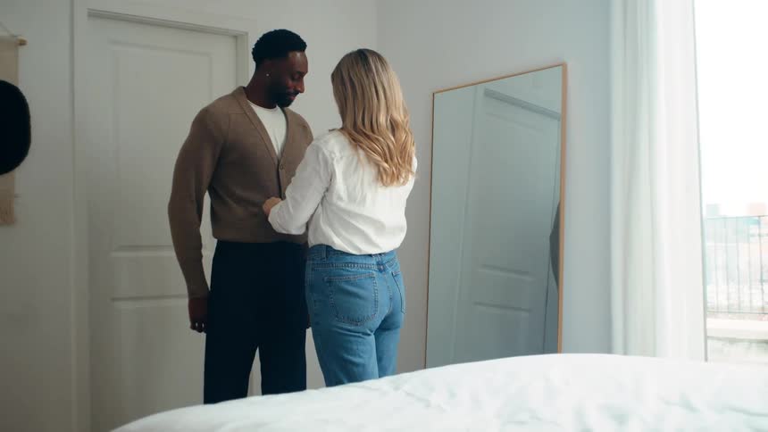 White blonde woman standing next to a black man arranging and tidying up his clothes in a bedroom indoors
