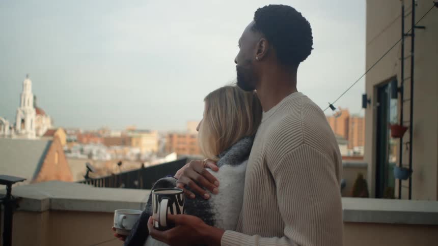 Blonde white woman wrapped in a blanket and black man standing up and hugging in a balcony looking at a suburban neighborhood around them outdoors during day time