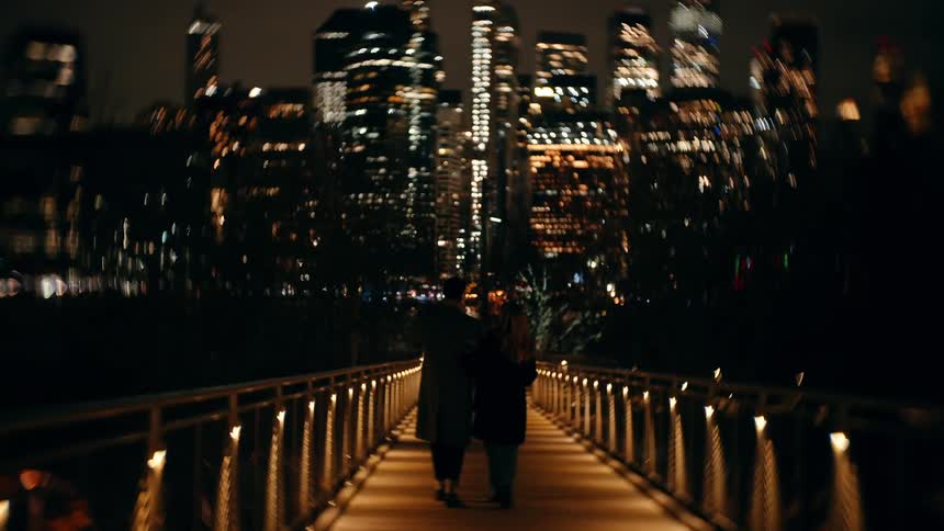 A couple walks on a welllit bridge at night with illuminated skyscrapers in the background
