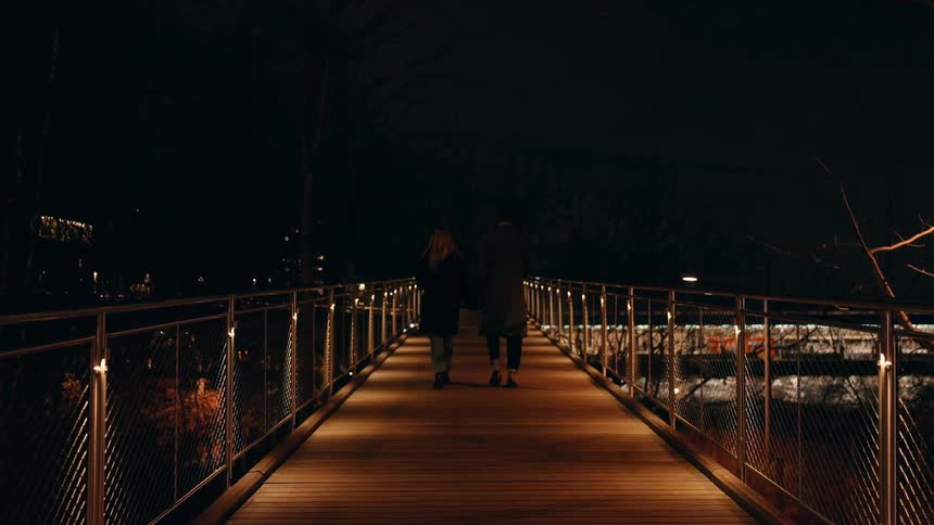 Two people are walking on a well lit wooden bridge at night over a pier in the city