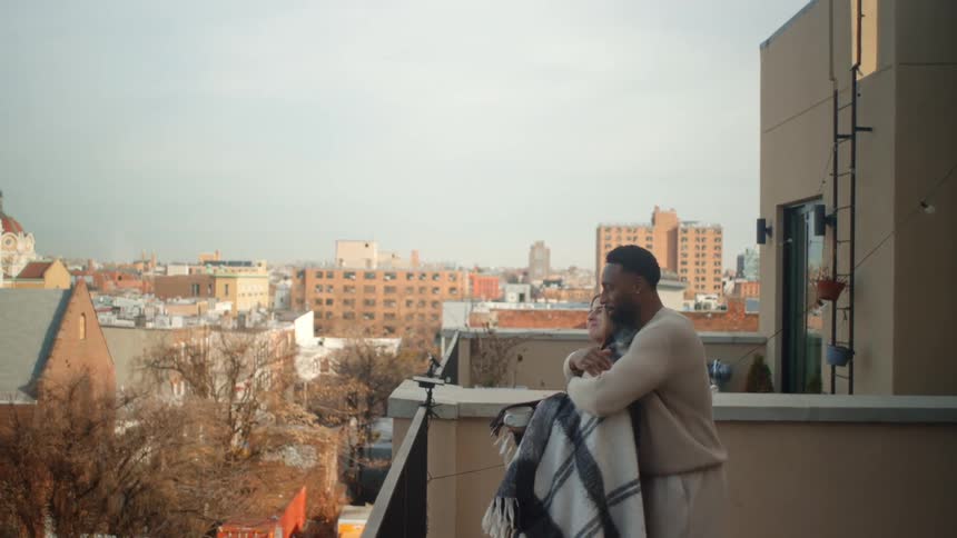 Black man covering a white blonde woman on a blanket then hugging her while both stand on a balcony and look at the view of a suburban area outdoors during day time