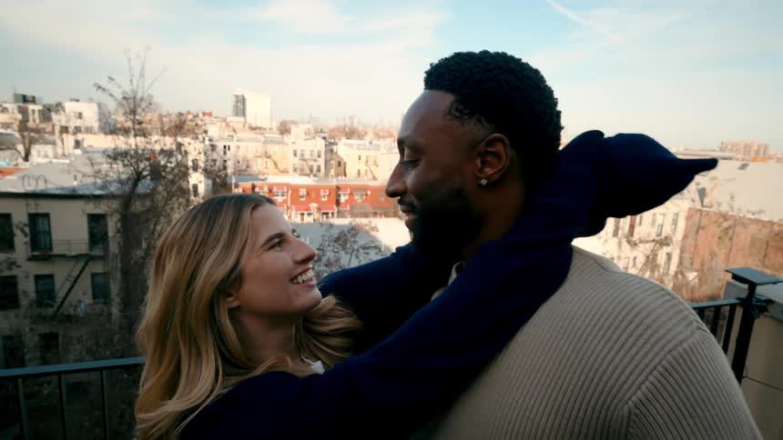 A young couple stands on a balcony embracing with a city under a blue sky in the background