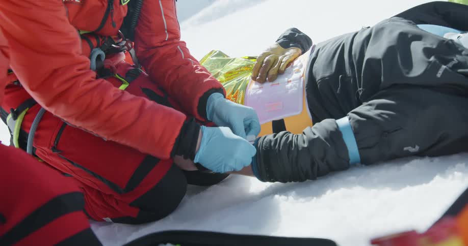 Doctors kneeling on the snowy ground outdoors rescuing a person lying down by applying bandage to their forearm during day time
