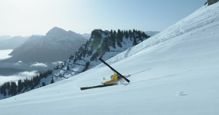 A skier in yellow gear falls on a snowy slope in the mountains