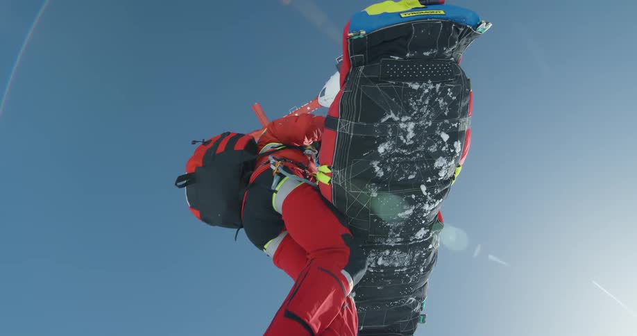 A person in red winter gear is airlifted with a large bag by a helicopter against a clear blue sky