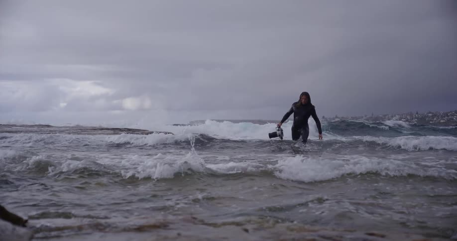 A person in a wetsuit walks through choppy ocean waves holding a camera on a cloudy day