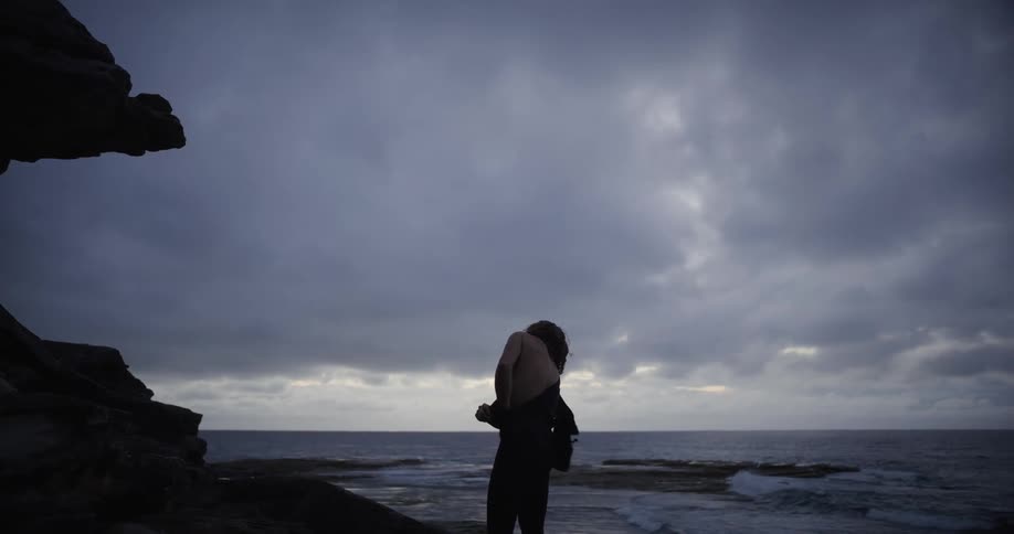 A silhouetted figure stands on a rocky shore by the ocean under a cloudy sky removing a dark garment