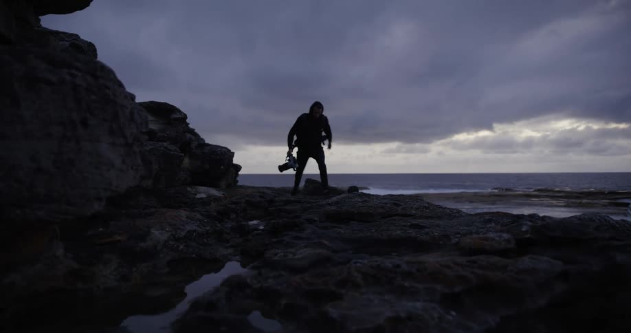 A silhouetted figure with a camera walks along a rocky coastline under a cloudy sky at dusk