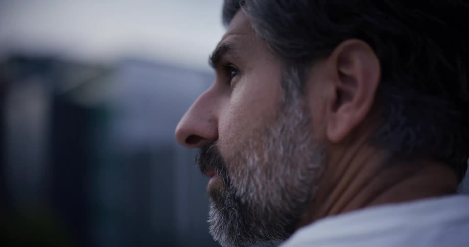 The profile of a man with a beard speckled with white standing outdoors against a blurred background