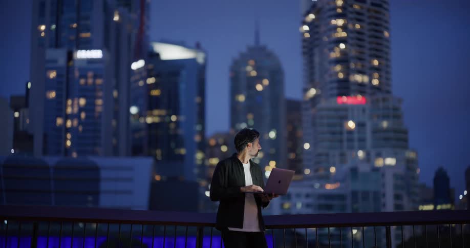 A bearded man in a black shirt over a white tshirt works on a laptop standing against a backdrop of illuminated skyscrapers at dusk