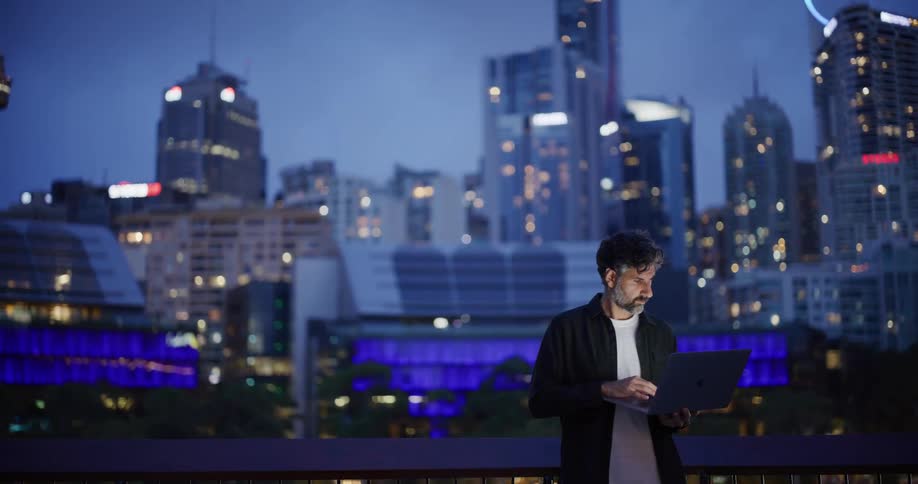 Bearded man in an outdoor environment typing on a laptop and in the background a metropolis illuminated at night
