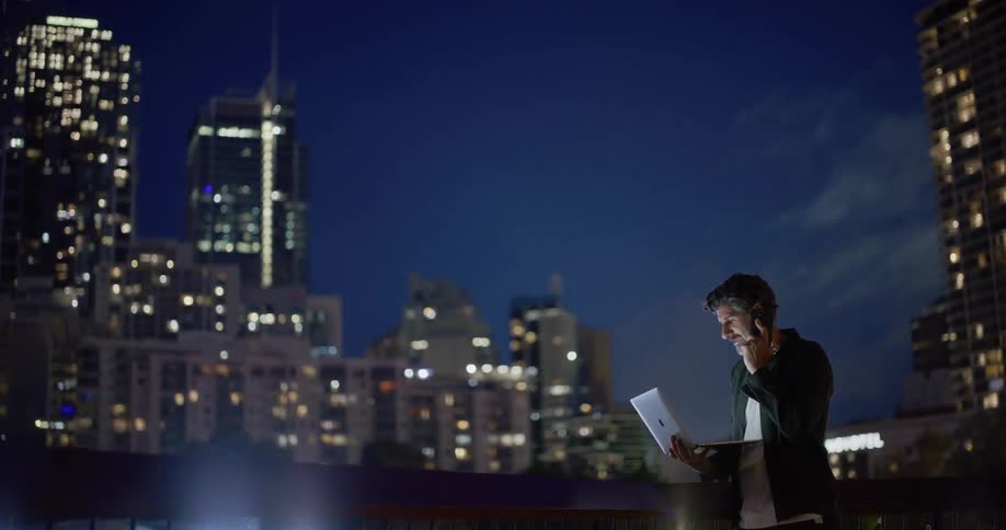 Older man holding an open laptop stands outdoors in the city at night and talks on his smartphone