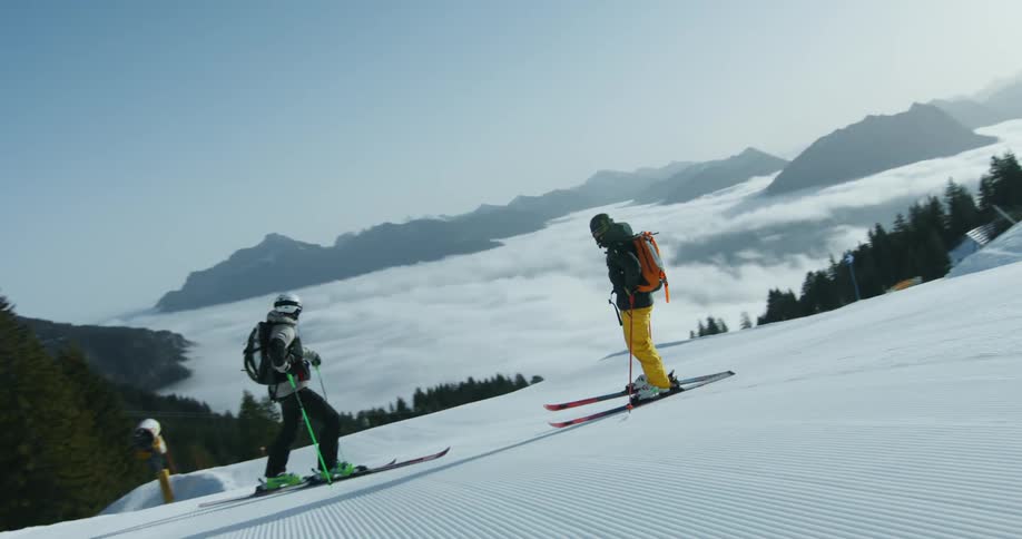 Two skiers stand on a groomed ski slope with a sea of clouds and mountains in the background