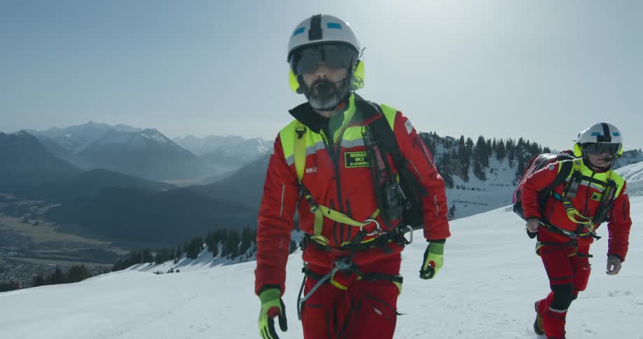 Rescue workers in red and yellow uniforms are climbing a snowy mountain with a view of distant mountain ranges and a valley below